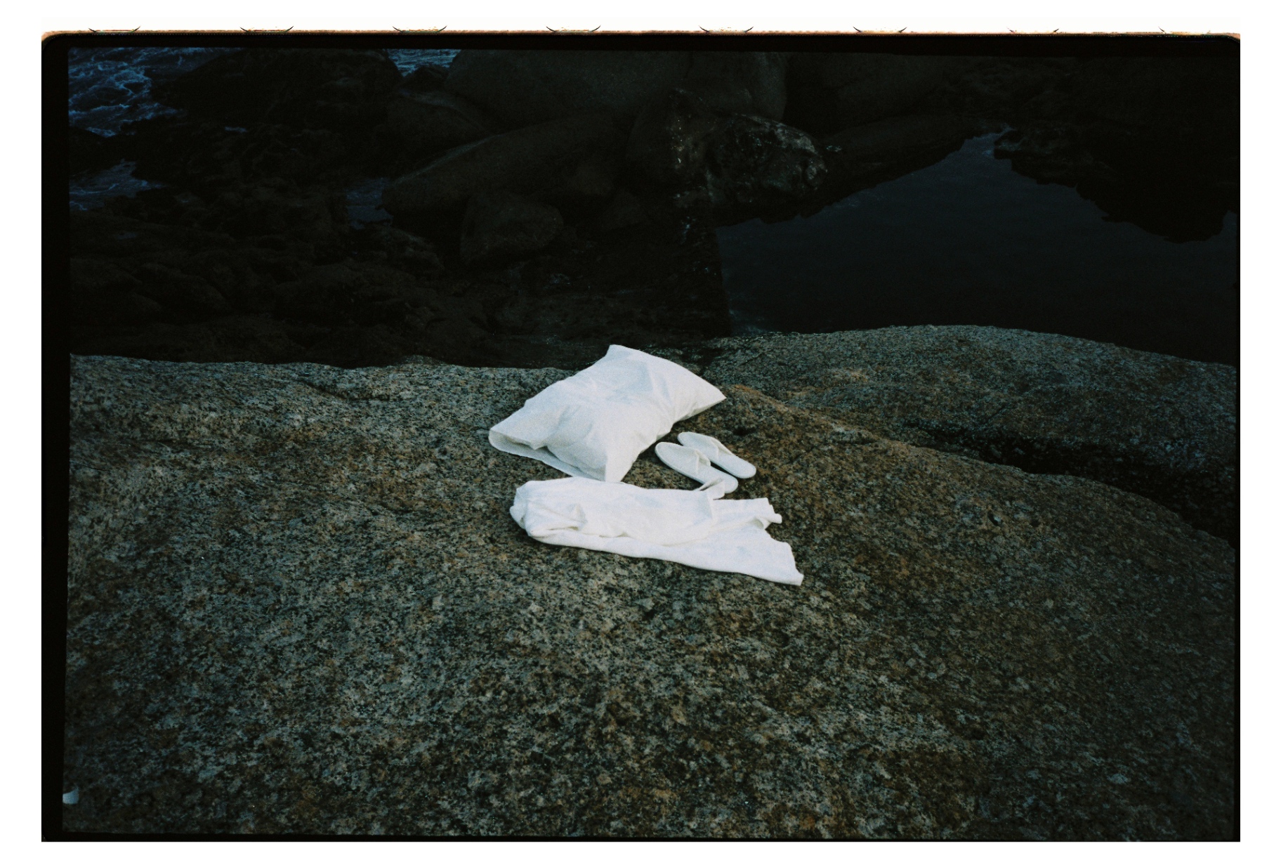 White Dover pillow, slippers, and folded robe on a dark coastal rock at dusk