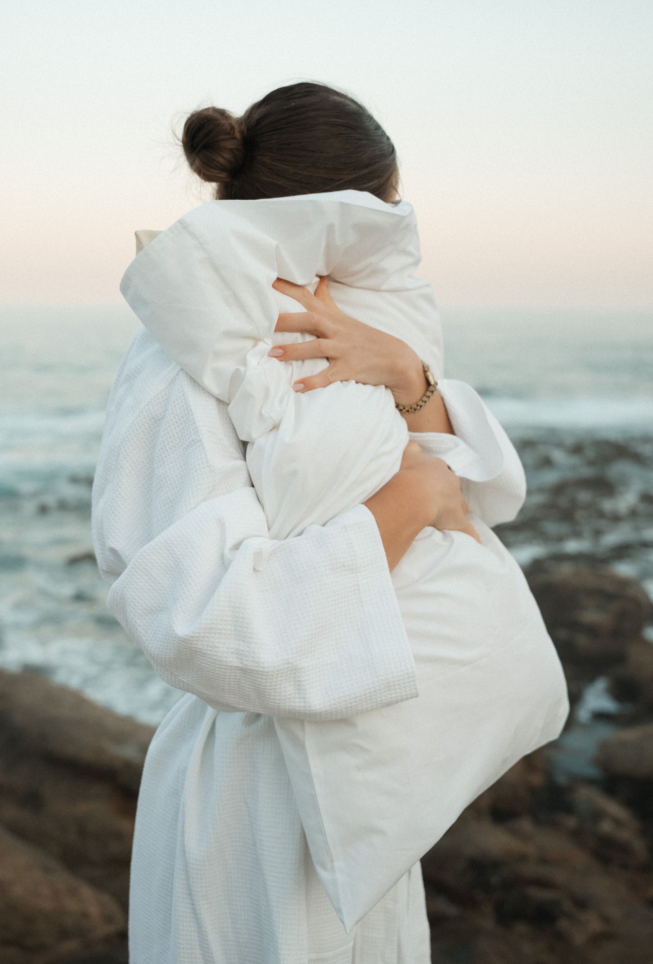 Woman hugging a crisp white Dover pillow on a rocky shoreline at dusk